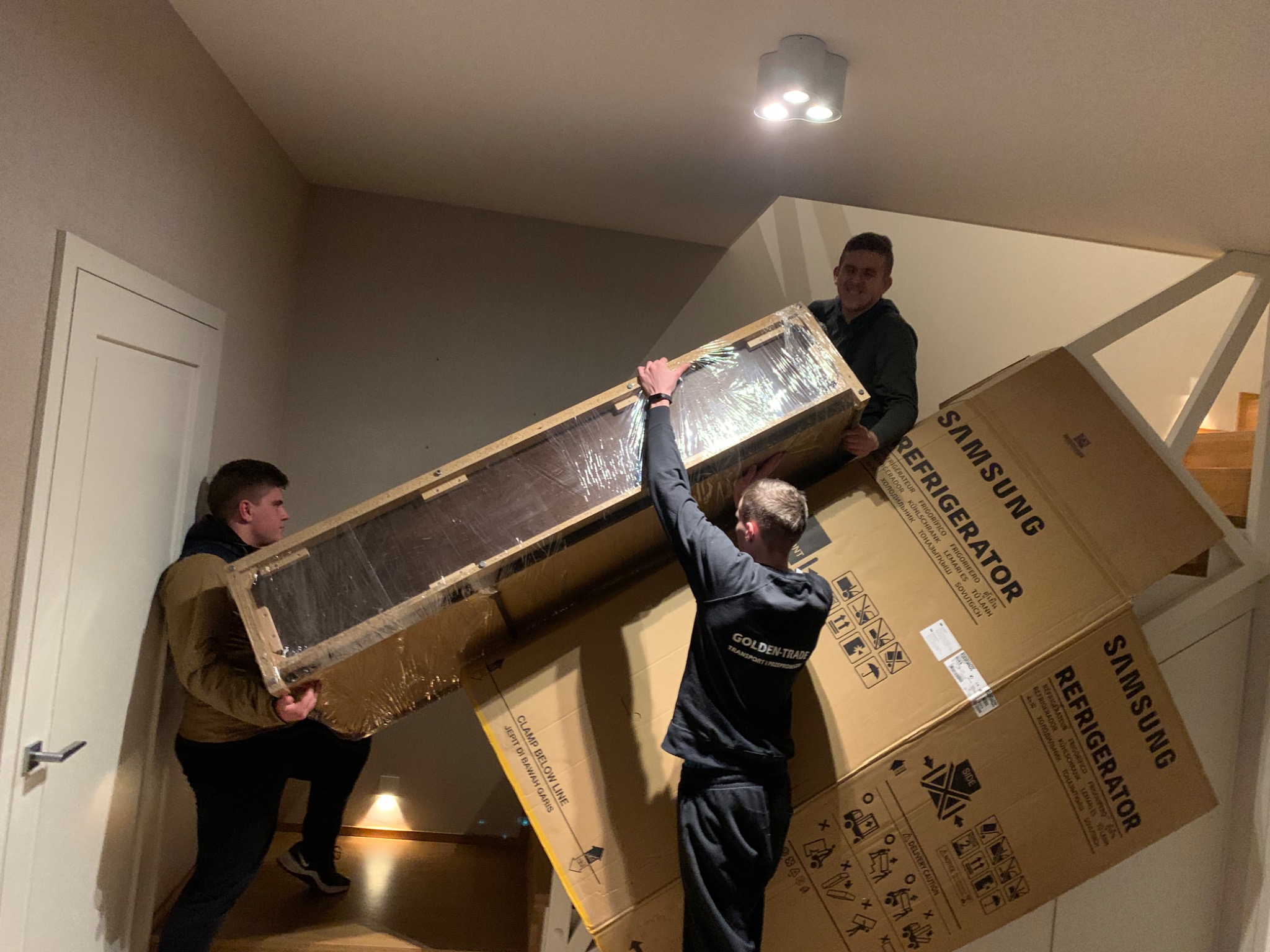 Trzech mężczyzn w czarnych ubraniach wnosi po schodach zapakowaną w folię i karton lodówkę marki Samsung, widoczne napisy ostrzegawcze na kartonie.