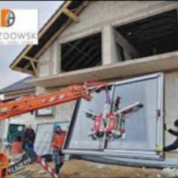 Grupa Perfekta Adam Drozdowski - Montaż dużego okna z użyciem dźwigu przyssawkowego na budowie domu jednorodzinnego. Widoczny pracownik w kamizelce odblaskowej i operator dźwigu. W tle surowy stan budynku z niezadaszonym dachem.