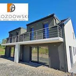 Grupa Perfekta Adam Drozdowski - Dwupiętrowy dom z płaskim dachem, szarą elewacją i dużymi przeszkleniami na parterze i piętrze, widoczne balustrady balkonów, logo firmy na górze zdjęcia.