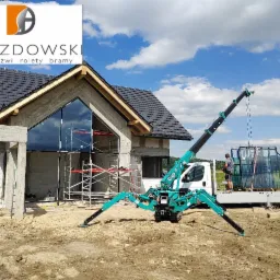 Montaż dużych okien za pomocą minidźwigu na placu budowy domu jednorodzinnego, widoczne rusztowania i logo firmy na budynku.