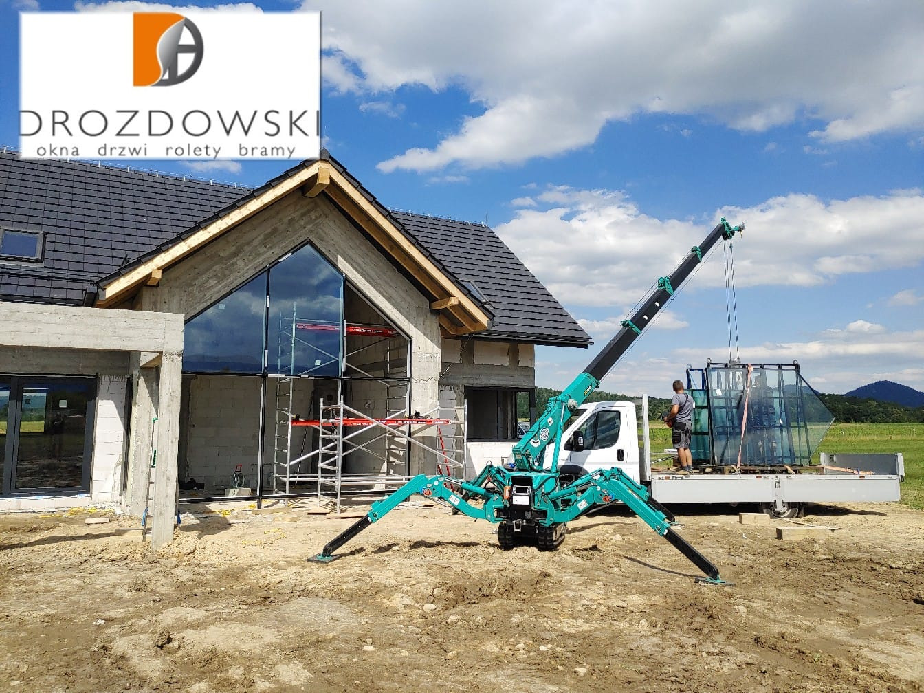 Montaż dużych okien za pomocą minidźwigu na placu budowy domu jednorodzinnego, widoczne rusztowania i logo firmy na budynku.