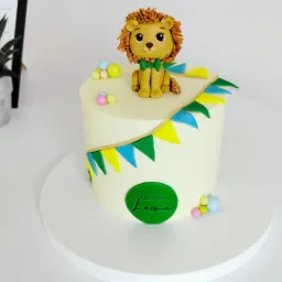 Tort na roczek z dekoracją w postaci lwa z masy cukrowej, otoczony girlandą z trójkątnych chorągiewek i kolorowymi kuleczkami, na białym tle.