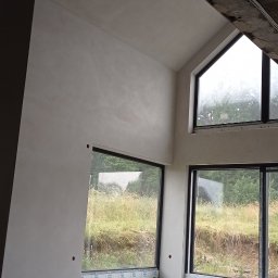 USŁUGI TYNKARSKO-BUDOWLANE - Surowe wnętrze z dużymi oknami i świeżo wykończonymi ścianami, widoczne ślady po szpachlowaniu i gładzi, betonowy sufit z instalacją elektryczną, widok na zewnątrz z zarośniętym terenem.