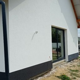 USŁUGI TYNKARSKO-BUDOWLANE - Nowoczesny dom jednorodzinny z jasną elewacją i ciemną podmurówką, widoczne okno z czarną ramą, rynna i fragment dachu, teren wokół domu w trakcie prac wykończeniowych.