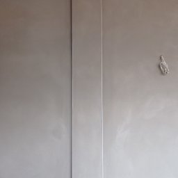 USŁUGI TYNKARSKO-BUDOWLANE - Ściana po nałożeniu szarej gładzi z pionową listwą przypodłogową, widoczne niedoskonałości i wystający kabel elektryczny.