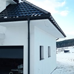 USŁUGI TYNKARSKO-BUDOWLANE - Boczny widok otynkowanego, białego domu z czarną dachówką i orynnowaniem w zimowej scenerii, z otwartym garażem i widokiem na zaśnieżone wzgórza w tle.