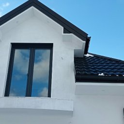 USŁUGI TYNKARSKO-BUDOWLANE - Fragment białej elewacji domu z czarnym dachem i oknem, odbijającym błękitne niebo, widoczne ślady po szpachlowaniu.