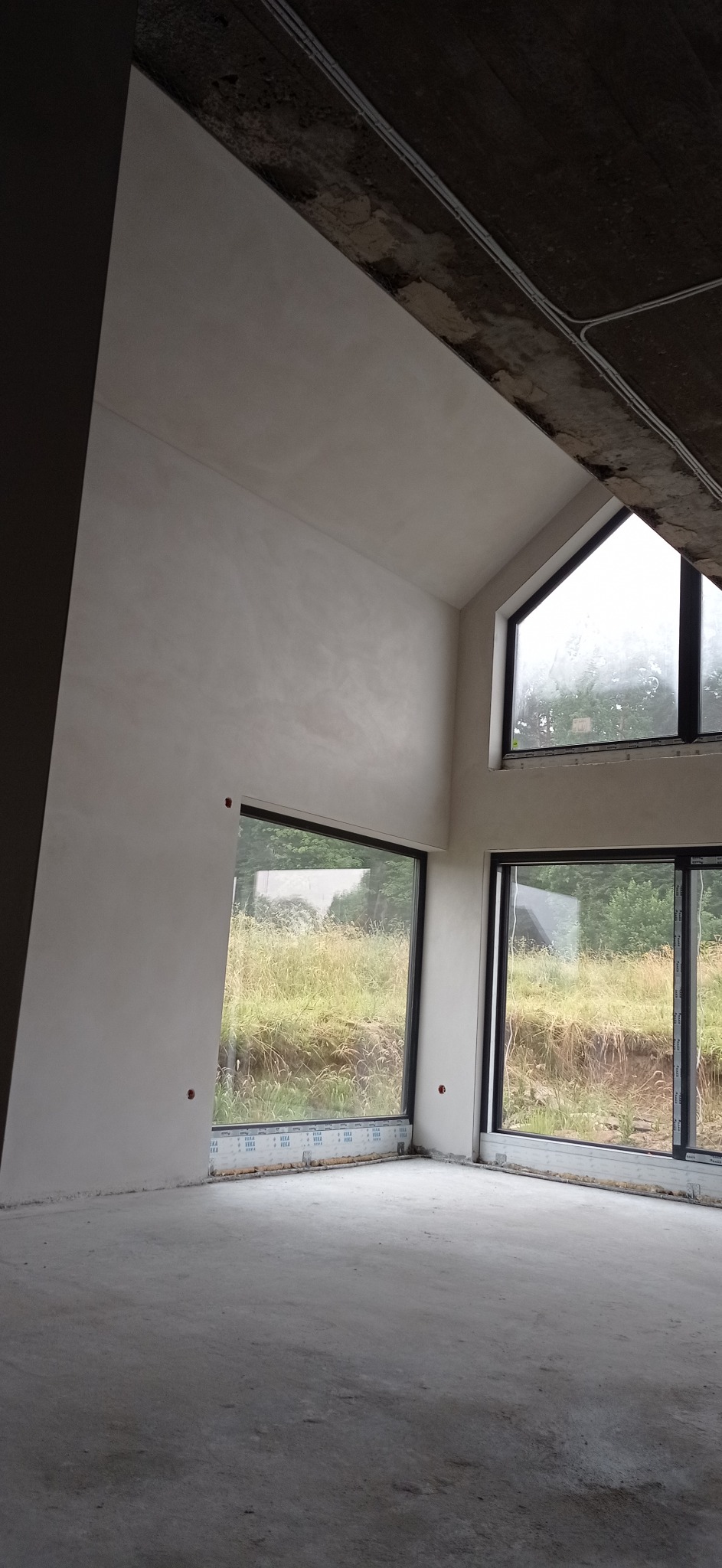 Surowe wnętrze z dużymi oknami i świeżo wykończonymi ścianami, widoczne ślady po szpachlowaniu i gładzi, betonowy sufit z instalacją elektryczną, widok na zewnątrz z zarośniętym terenem.