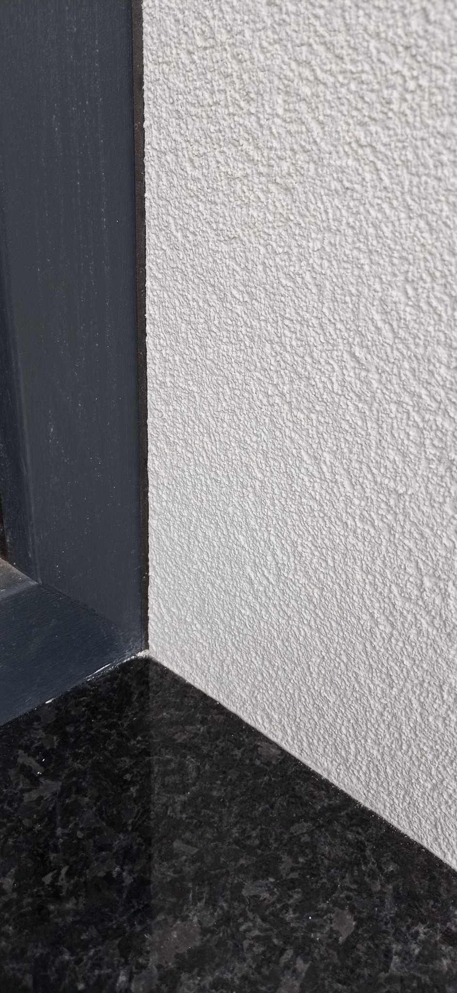 Szara ściana z tynkiem barankiem, ciemna rama okienna i czarny granitowy parapet.