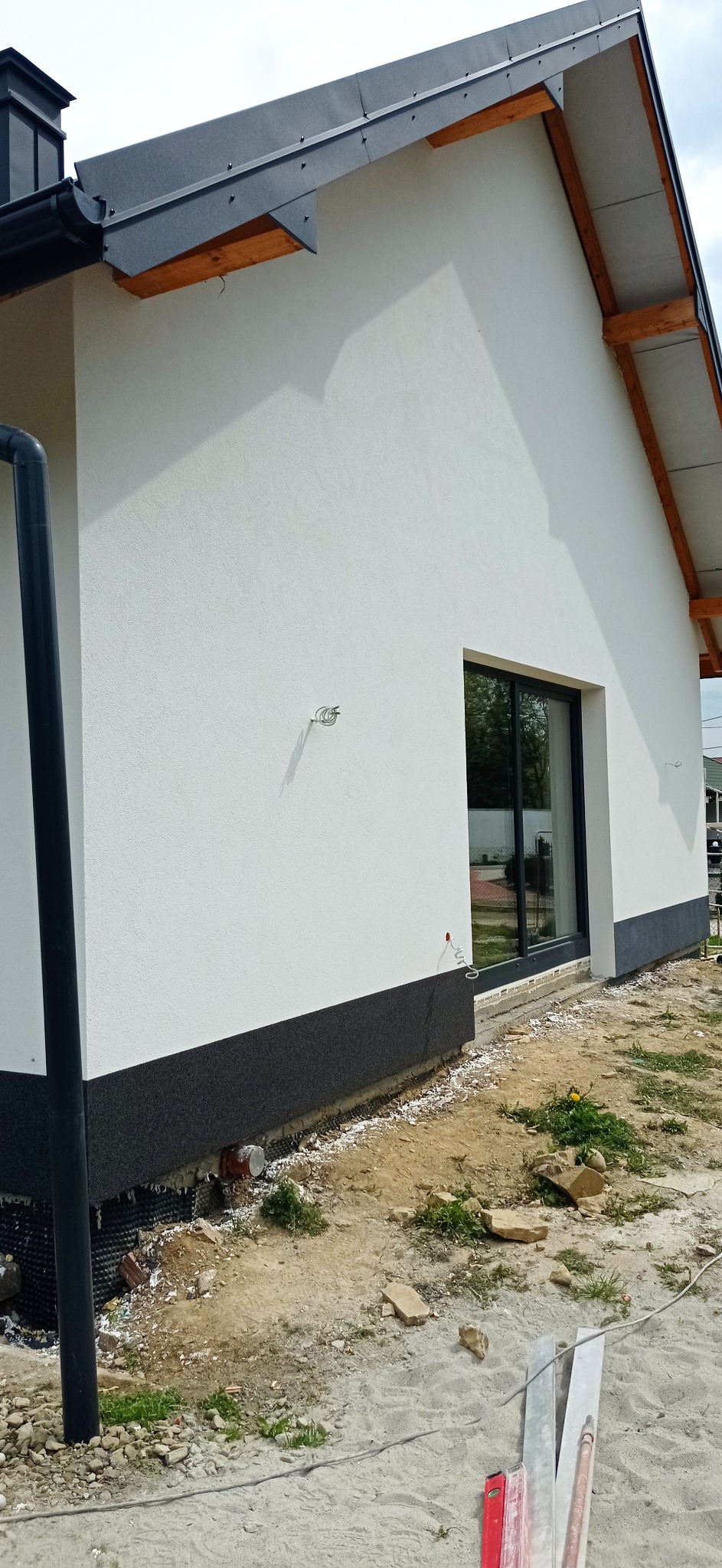 Nowoczesny dom jednorodzinny z jasną elewacją i ciemną podmurówką, widoczne okno z czarną ramą, rynna i fragment dachu, teren wokół domu w trakcie prac wykończeniowych.