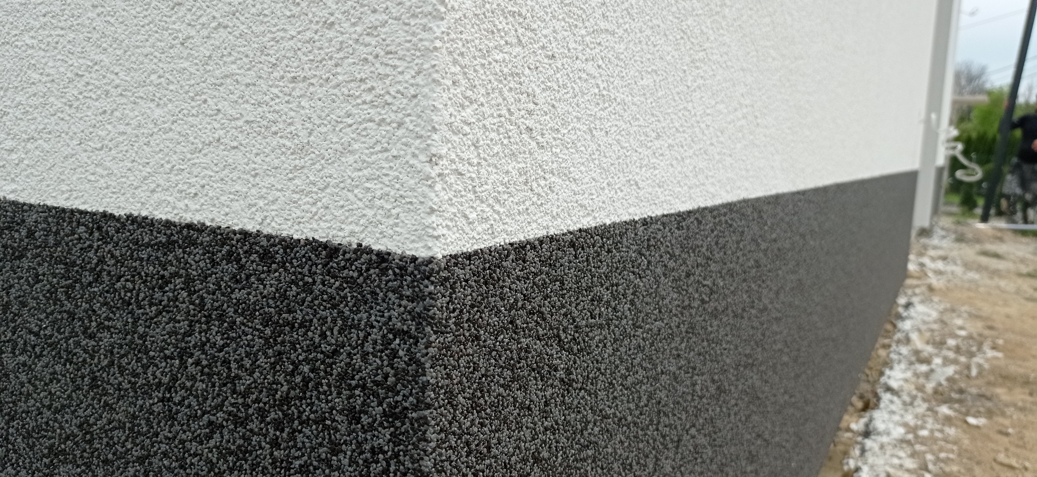 Fragment elewacji budynku z wyraźnie oddzieloną, dwukolorową strukturą tynku: górna część biała, dolna ciemnoszara, z widoczną fakturą baranka, zarys narożnika i fragment otoczenia zewnętrznego.