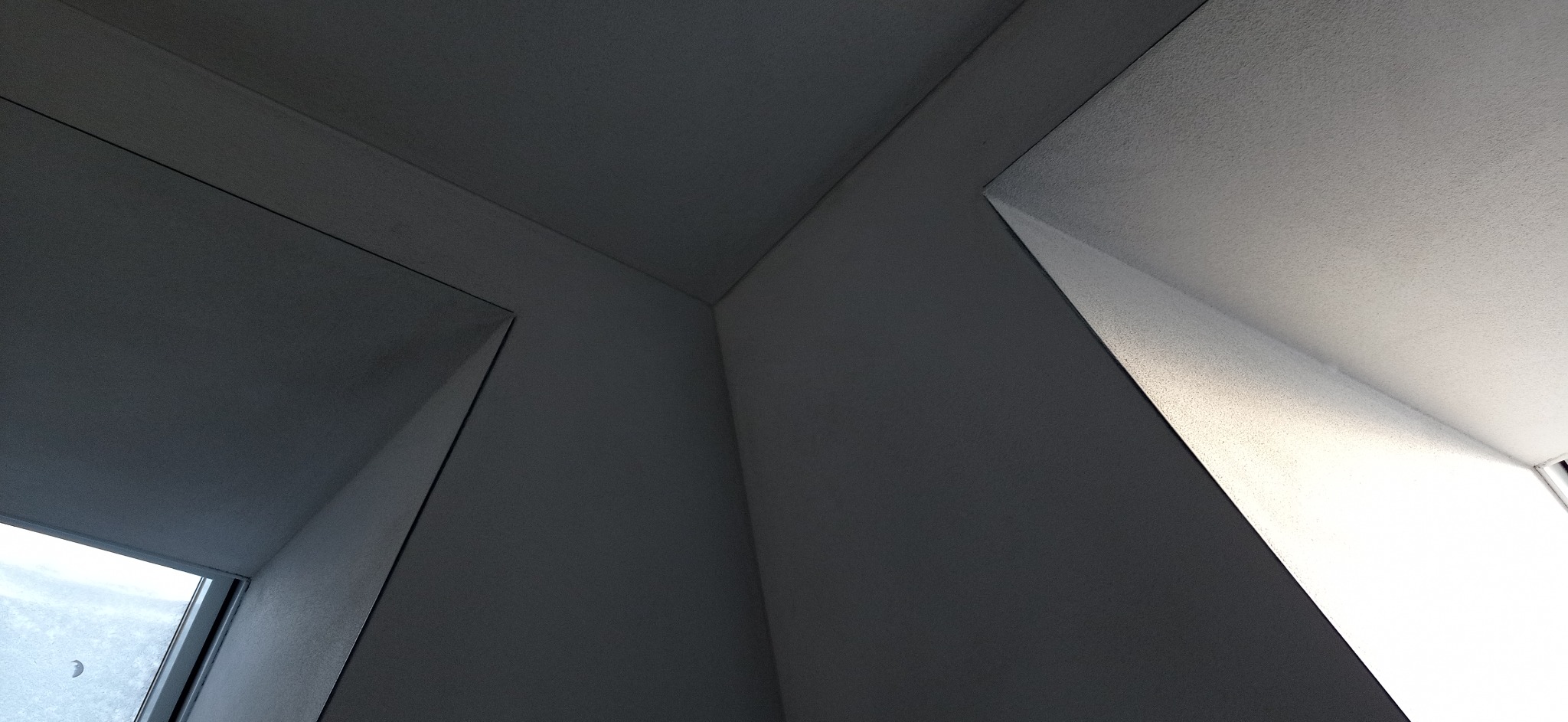 Wnętrze pomieszczenia z surowymi ścianami i sufitem, widok z dołu na geometryczne połączenia powierzchni, światło dzienne wpadające przez okno.