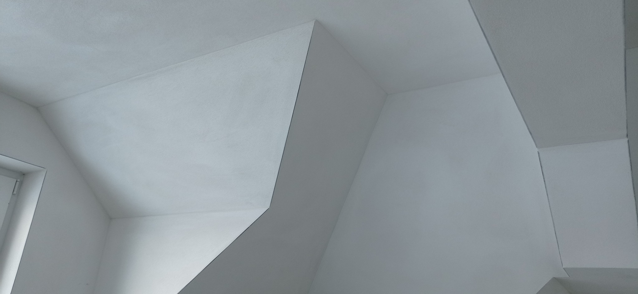 Wnętrze poddasza po świeżo wykonanych pracach wykończeniowych, widoczne gładkie, białe ściany i sufit z nietypowymi skosami i załamaniami, delikatne światło wpadające przez okno po lewej stronie.