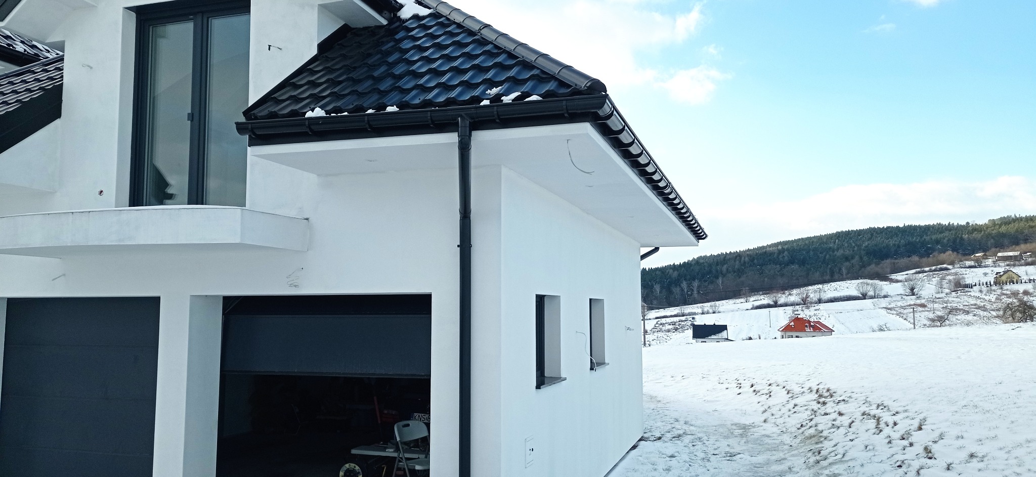 Boczny widok otynkowanego, białego domu z czarną dachówką i orynnowaniem w zimowej scenerii, z otwartym garażem i widokiem na zaśnieżone wzgórza w tle.