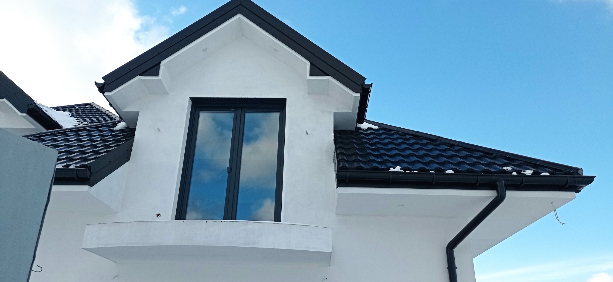 Fragment białej elewacji domu z czarnym dachem i oknem, odbijającym błękitne niebo, widoczne ślady po szpachlowaniu.