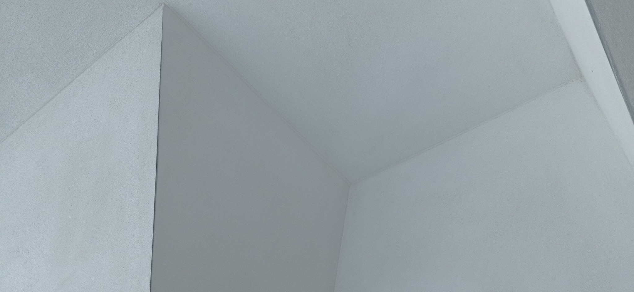 Wnętrze pomieszczenia z gładkimi, pomalowanymi na biało ścianami i sufitem, widok na narożnik z delikatnym oświetleniem padającym z góry.