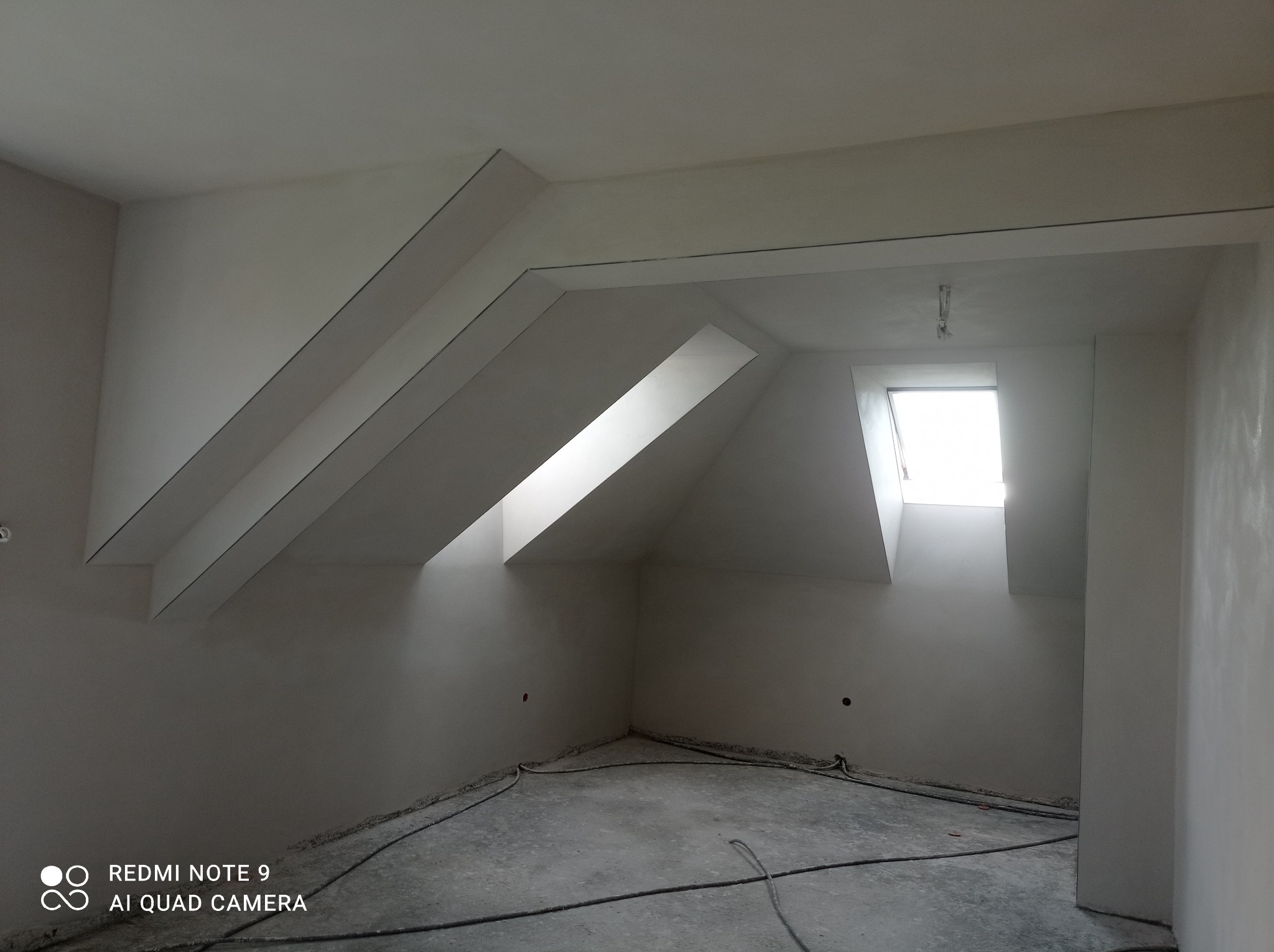 Poddasze po gipsowaniu ścian i sufitu, widoczne okno dachowe i kable na podłodze, ściany przygotowane do malowania.