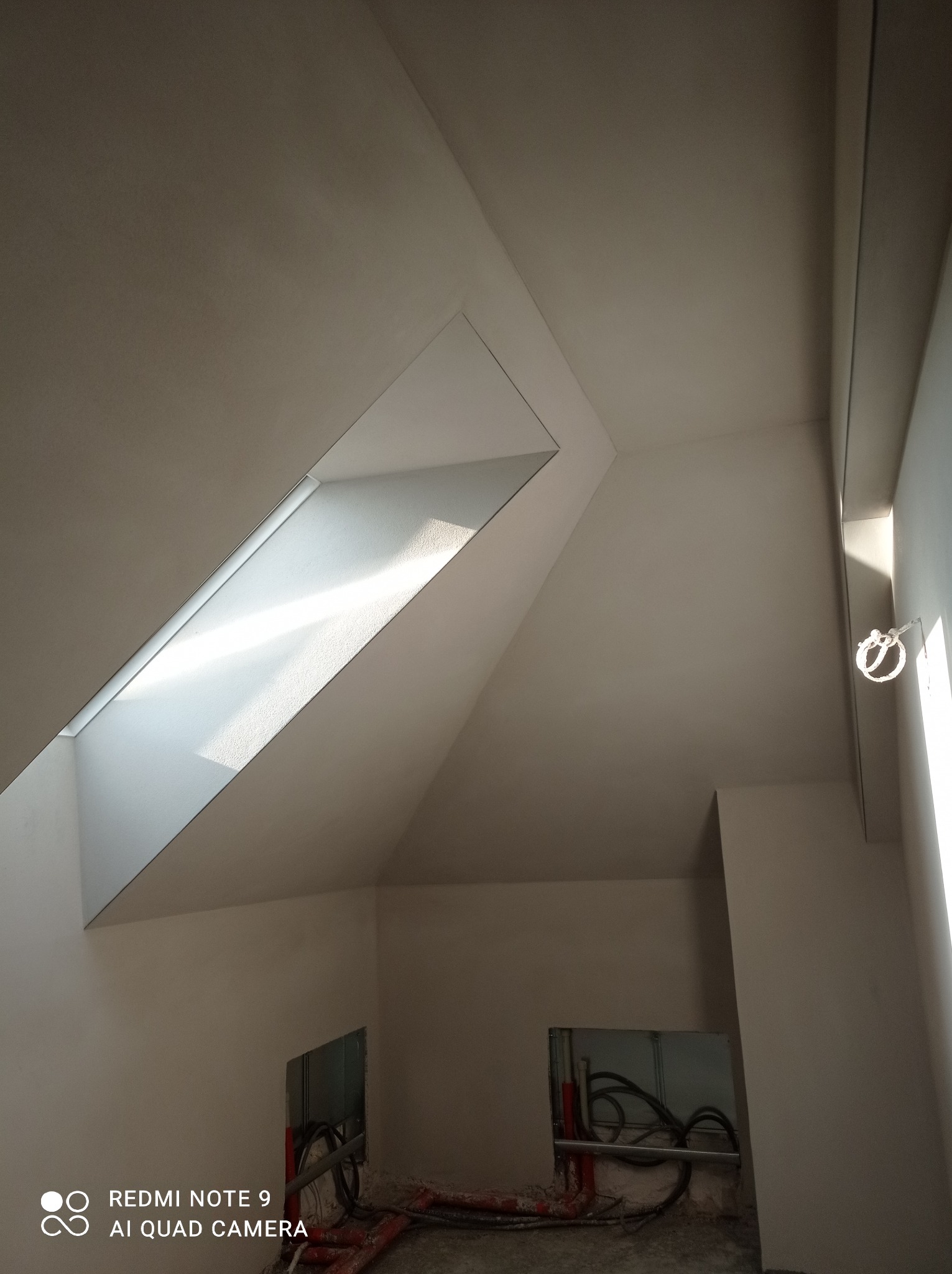 Wnętrze w trakcie prac wykończeniowych: ściany przygotowane pod malowanie, widoczne instalacje elektryczne i hydrauliczne, okno dachowe wpuszczające jasne światło.