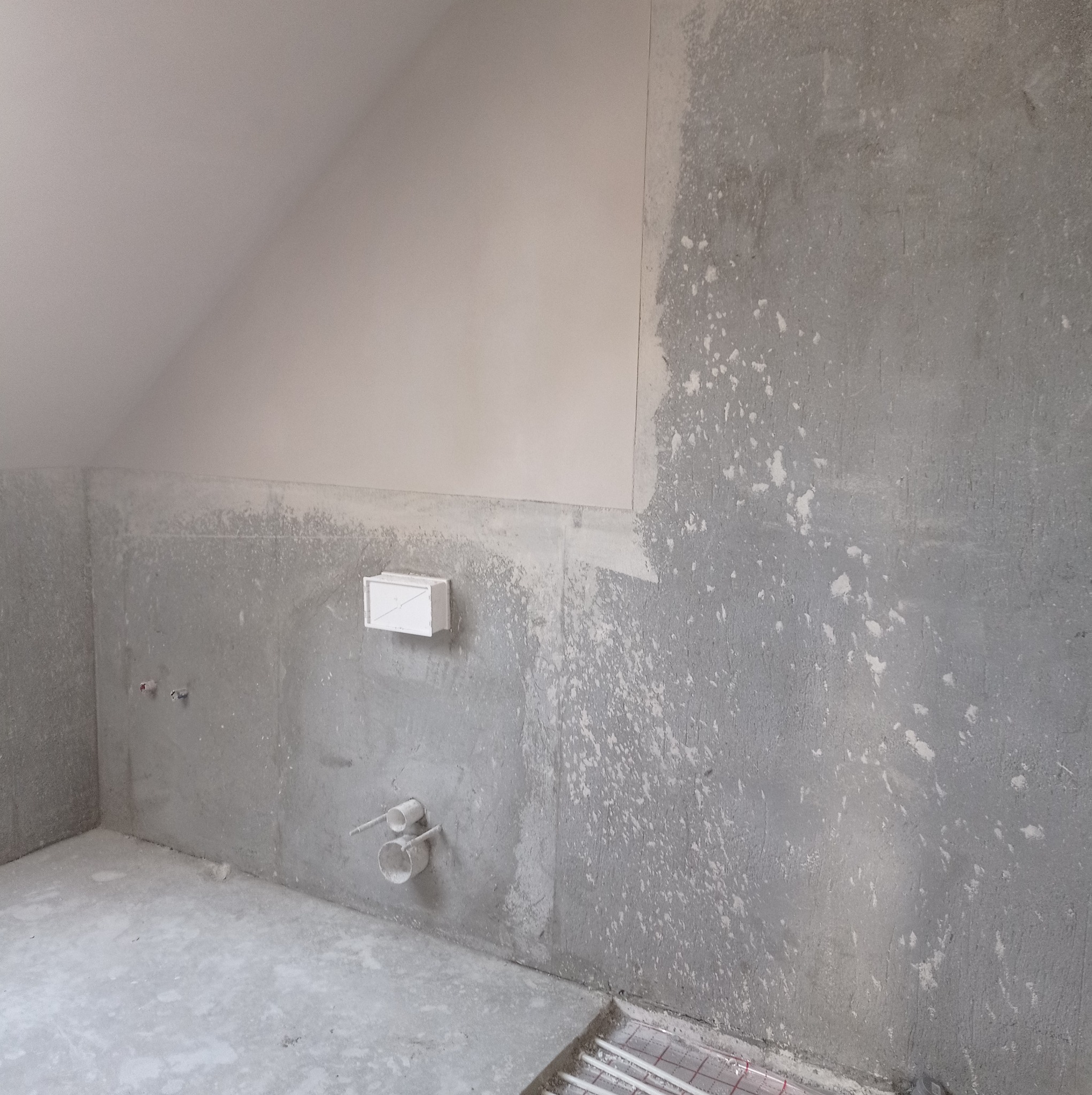 Wnętrze w trakcie remontu: ściana częściowo pokryta gładzią, widoczne instalacje hydrauliczne i ogrzewanie podłogowe, surowy stan wykończenia.