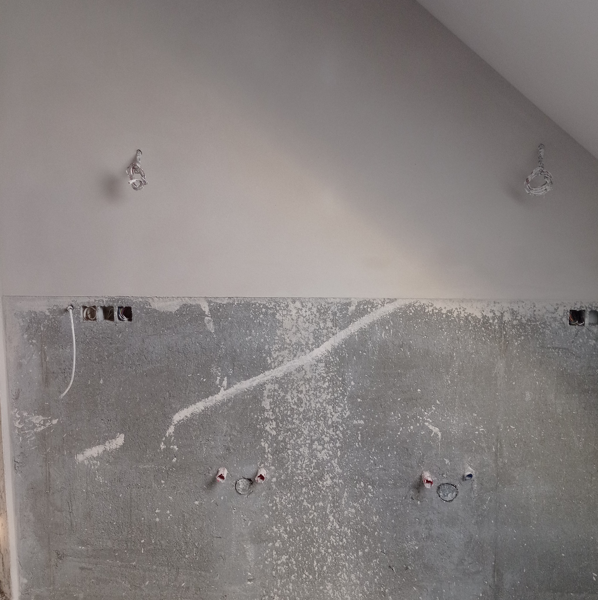 Surowa ściana z widocznymi przewodami elektrycznymi i rurami, przygotowywana do dalszych prac wykończeniowych, z fragmentem skośnego sufitu i wystającymi przewodami oświetleniowymi.