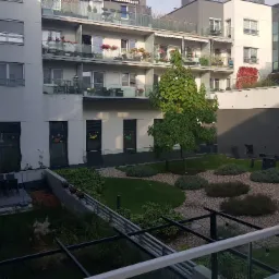 Wewnętrzny dziedziniec nowoczesnego budynku mieszkalnego z trawnikiem, krzewami i drzewem, widok z wyższego piętra przez metalową balustradę balkonu.