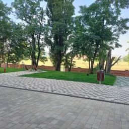 Ułożona kostka brukowa prowadząca do trawnika z drzewami, czerwonym hydrantem i dwoma leżakami na tle ceglanego murku i pola.