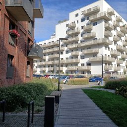 ŁADNY DOM - Nowoczesne osiedle mieszkaniowe z ceglanym i białym budynkiem, kostką brukową, zielenią i miejscami parkingowymi w słoneczny dzień.