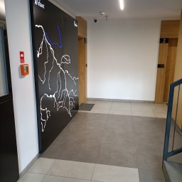 ŁADNY DOM - Korytarz w nowoczesnym budynku z tablicą informacyjną przedstawiającą schemat rzeki Widawy w białym i niebieskim kolorze, widoczne drzwi do mieszkań i fragment schodów.