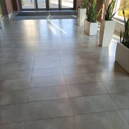 ŁADNY DOM - Błyszcząca, szara posadzka w holu biurowca, z rzędem wysokich, białych donic z zielonymi roślinami wzdłuż ściany i przeszklonym wejściem w tle.