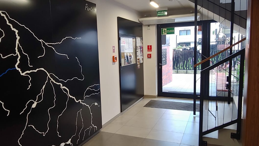 Wnętrze klatki schodowej w budynku mieszkalnym z czarną ścianą ozdobioną białą mapą rzeki, widoczne schody z czarnymi balustradami, drzwi wejściowe z oknem i tablicą informacyjną.
