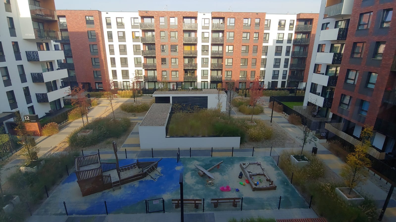 Widok z góry na nowoczesne osiedle z placem zabaw w kształcie statku i samolotu, otoczone budynkami z ceglanymi i białymi elewacjami oraz ozdobnymi trawami.