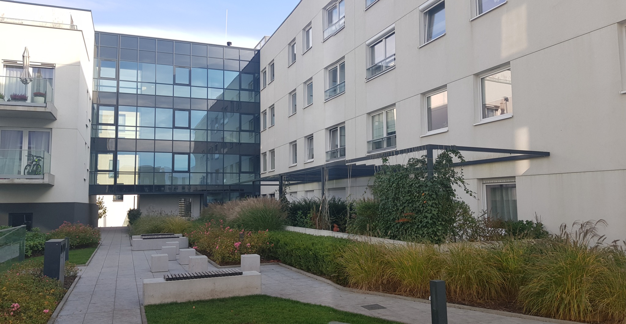 Nowoczesny dziedziniec pomiędzy budynkami z biurami i mieszkaniami, z elementami małej architektury, zadbaną zielenią i trawami ozdobnymi.