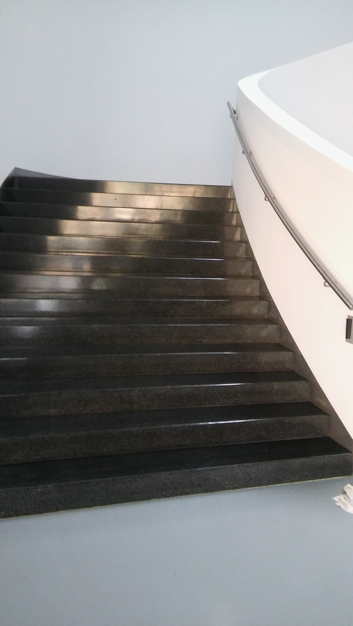 Ciemne, granitowe schody w budynku biurowym, z jasnymi ścianami i metalową poręczą, widoczne z dołu, z lekkim odblaskiem światła na stopniach.