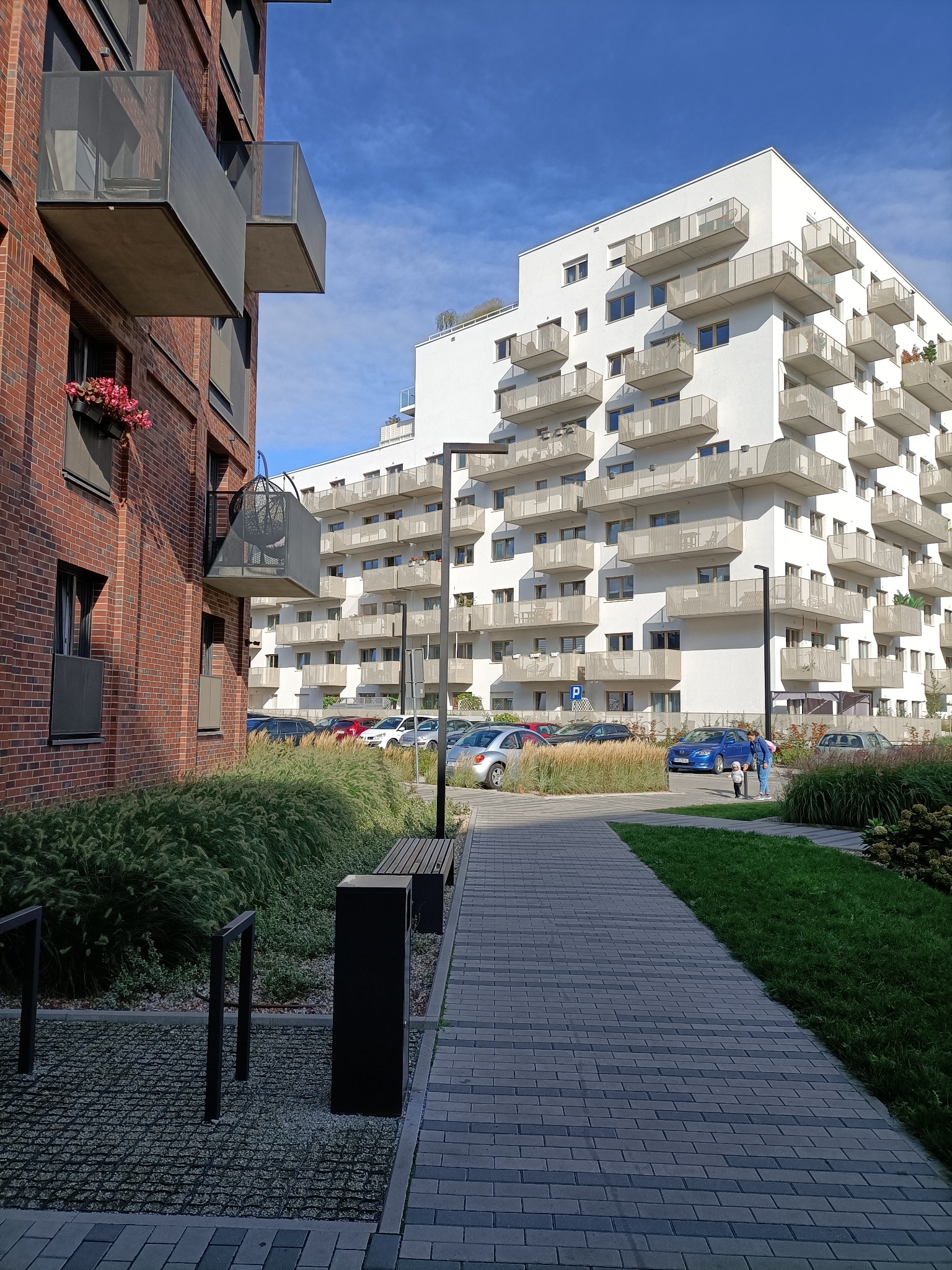 Nowoczesne osiedle mieszkaniowe z ceglanym i białym budynkiem, kostką brukową, zielenią i miejscami parkingowymi w słoneczny dzień.