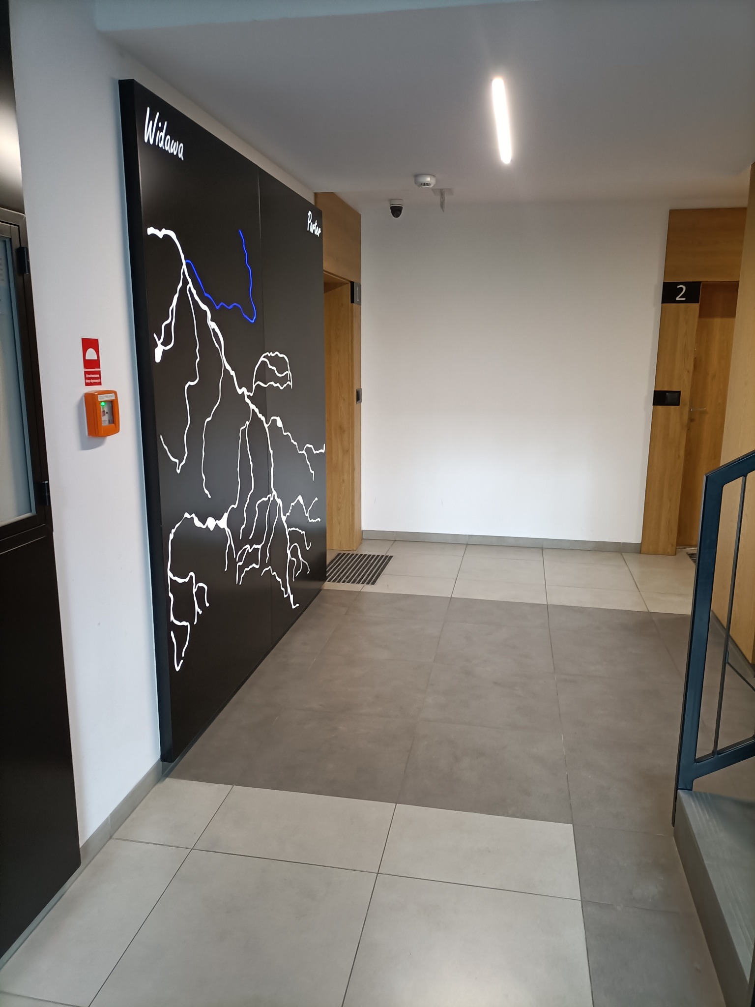 Korytarz w nowoczesnym budynku z tablicą informacyjną przedstawiającą schemat rzeki Widawy w białym i niebieskim kolorze, widoczne drzwi do mieszkań i fragment schodów.