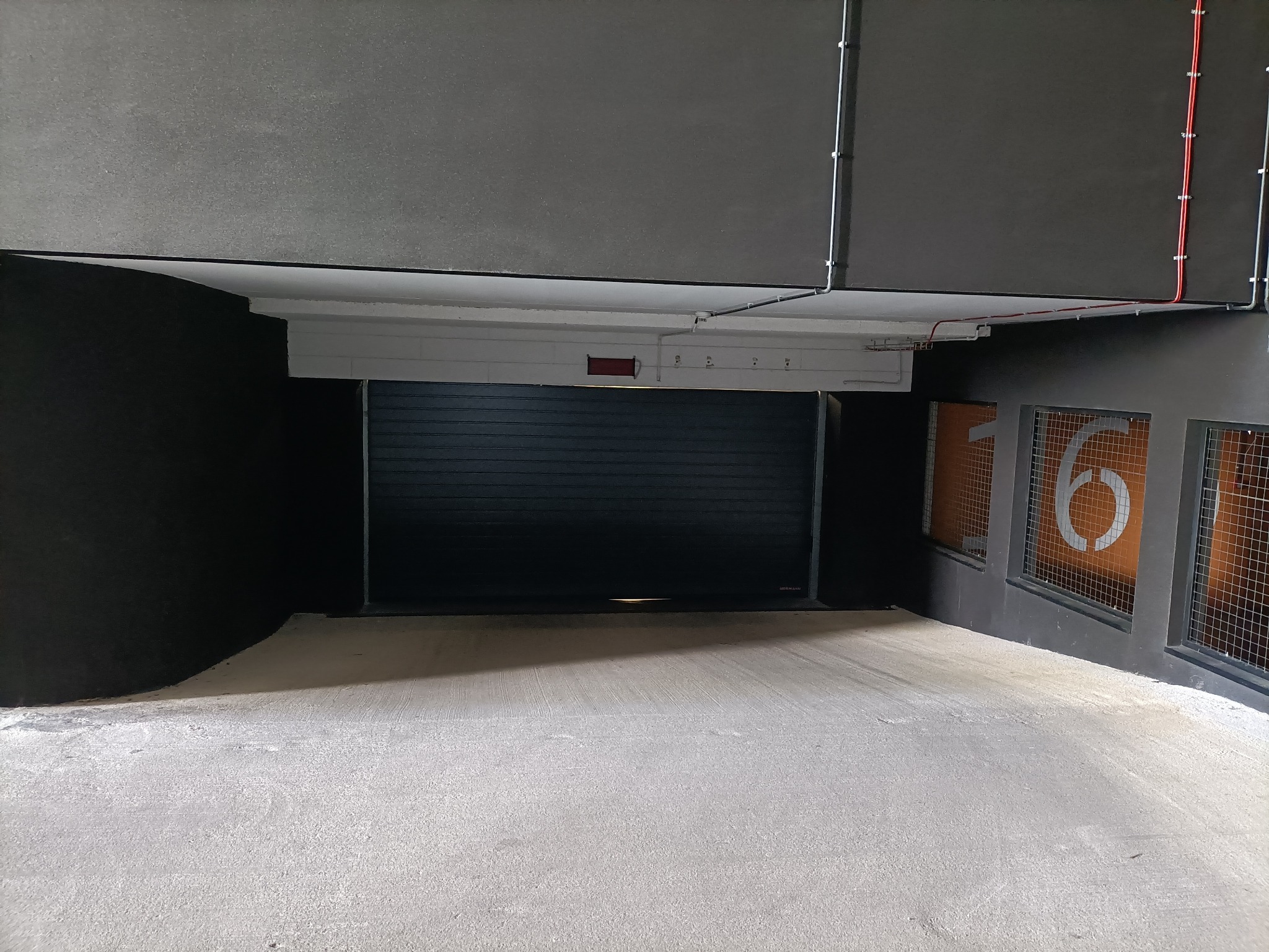 Wjazd do garażu podziemnego z zamkniętą, metalową bramą rolowaną, widoczne numery stanowisk parkingowych za metalową siatką, szara betonowa posadzka i ciemne ściany.