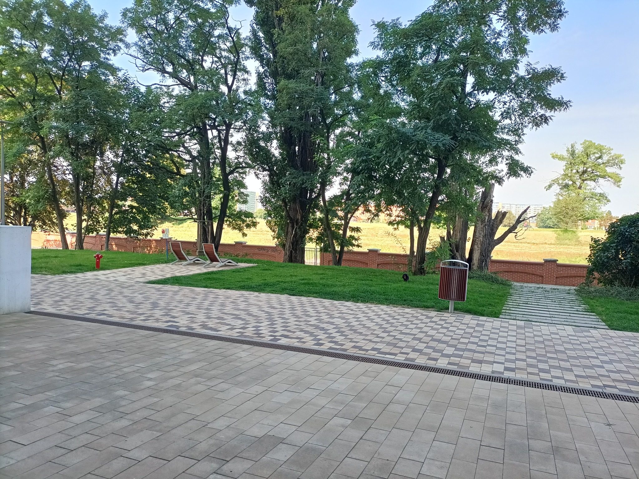 Ułożona kostka brukowa w szarych odcieniach tworząca plac przed budynkiem, z widocznym systemem odwadniającym, przechodząca w ścieżkę i trawnik z drzewami, z elementami małej architektury jak kosz...