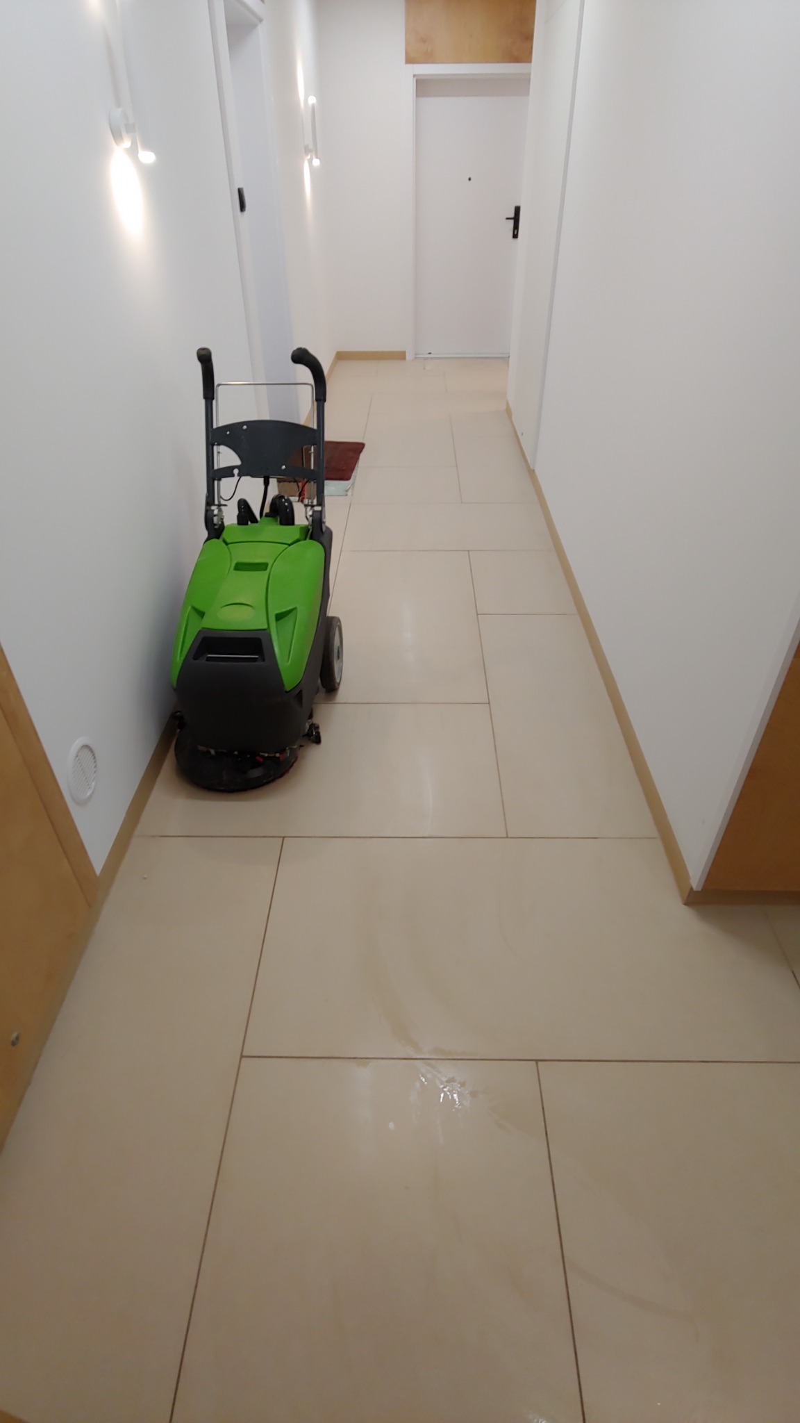 Profesjonalna, zielona maszyna do czyszczenia podłóg stoi na jasnych, ceramicznych płytkach w korytarzu biurowym. Widoczne ślady wilgoci na podłodze po czyszczeniu.