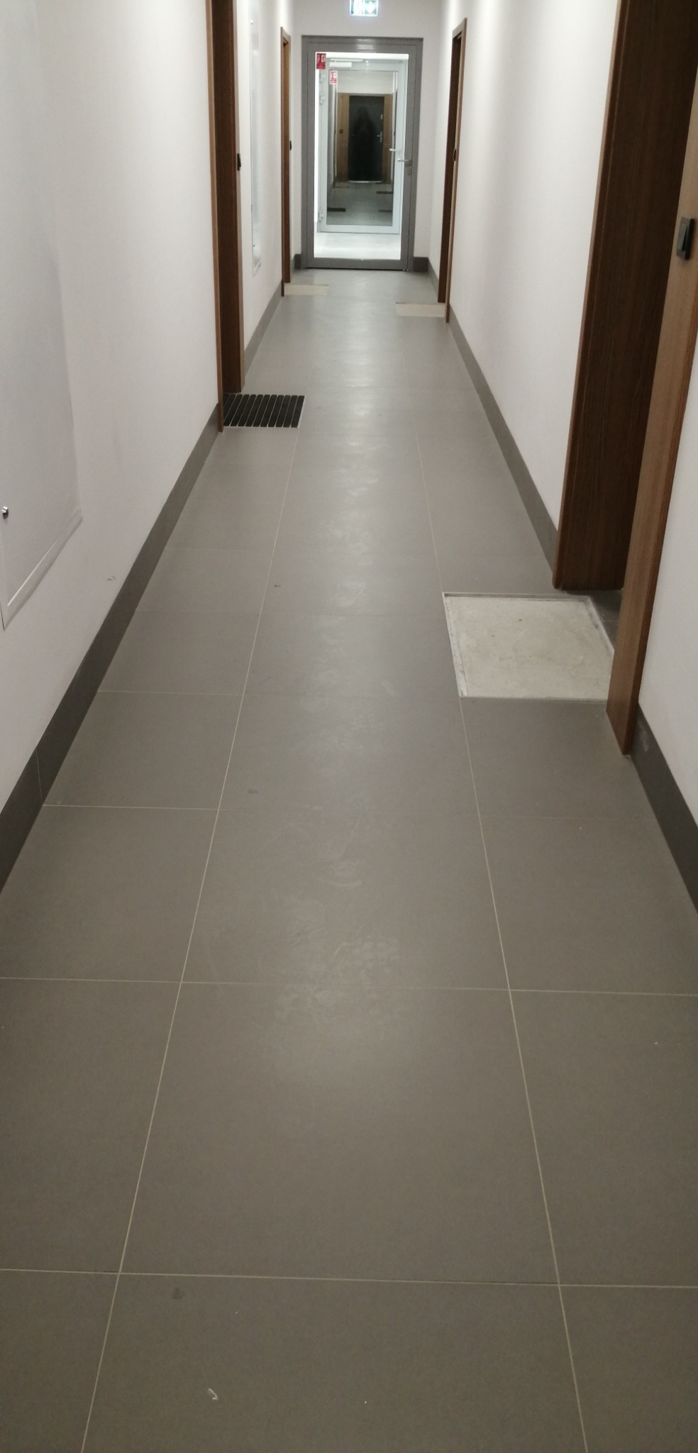 Długi korytarz biurowy z szarą, kwadratową podłogą wyłożoną płytkami, białymi ścianami i ciemnymi framugami drzwi. W oddali widoczne okno lub drzwi na końcu korytarza.