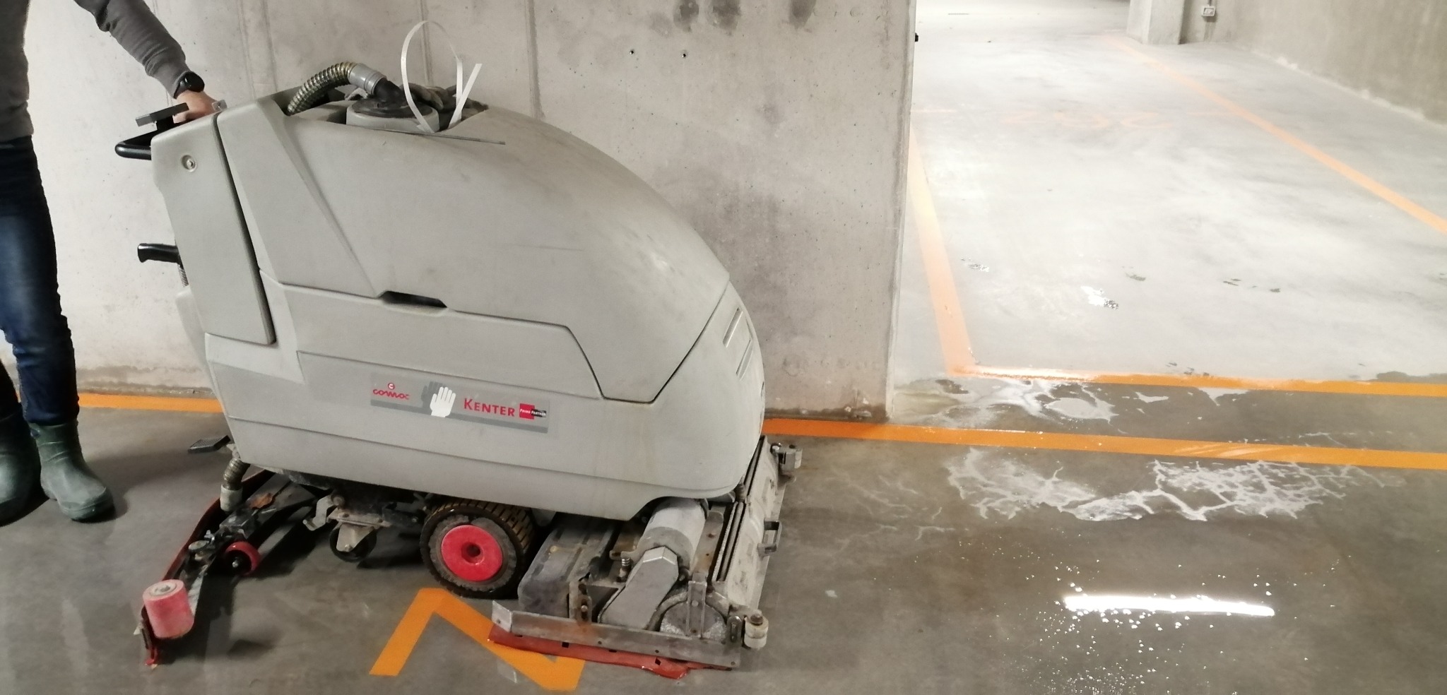 Operator prowadzi maszynę do szorowania podłóg w garażu podziemnym, widoczne mokre ślady po czyszczeniu i pomarańczowe linie wyznaczające miejsca parkingowe.