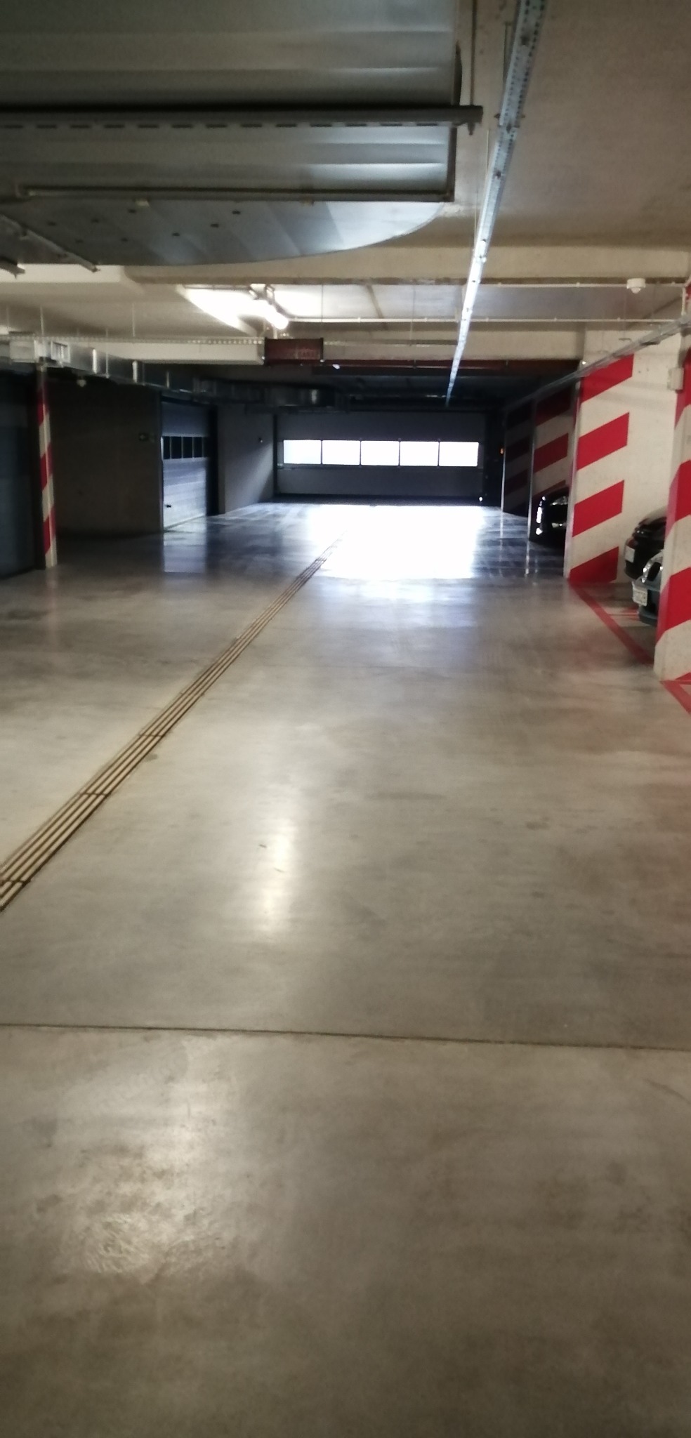 Wnętrze parkingu podziemnego z betonową posadzką, oświetleniem sufitowym i kolumnami oznaczonymi czerwono-białymi pasami, z widocznymi elementami systemu nawigacji dla osób niewidomych na posadzce.