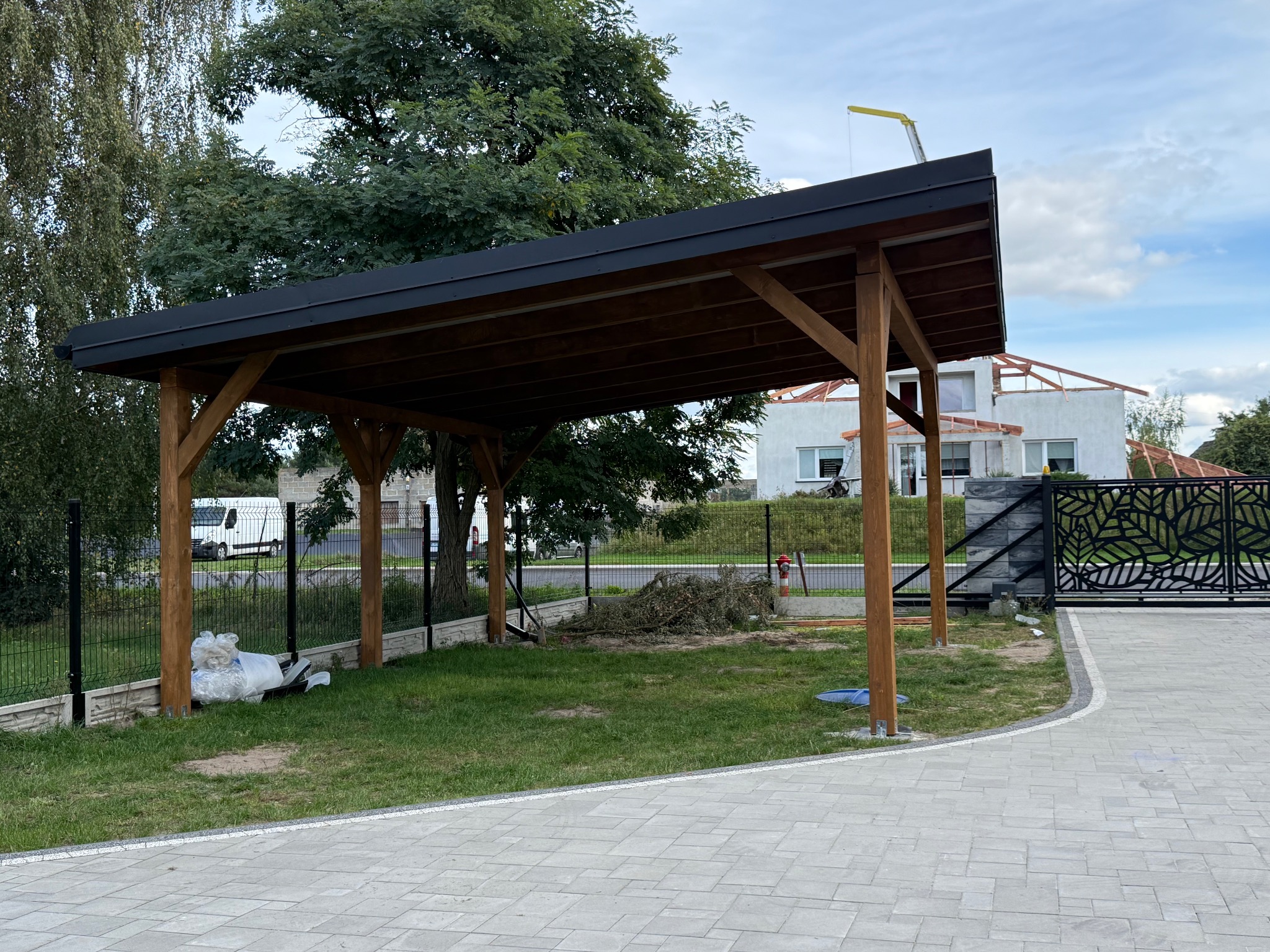 Zadaszenie tarasu z drewnianą konstrukcją i czarnym dachem, widoczne z zewnątrz. W tle dom w trakcie budowy i zielone drzewa. Ujęcie z poziomu kostki brukowej.