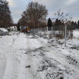 PREMIUMSTAL SEBASTIAN PRZYBYSZ - Montaż siatki ogrodzeniowej na metalowych słupkach w zimowej scenerii, widoczny mały traktor i dwóch pracowników w oddali.