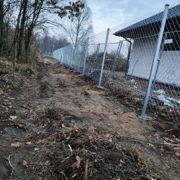 PREMIUMSTAL SEBASTIAN PRZYBYSZ - Długa linia siatki ogrodzeniowej z metalowymi słupkami, biegnąca wzdłuż nierównego terenu porośniętego drzewami i liśćmi, z fragmentem budynku w tle.