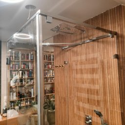 PREMIUMSTAL SEBASTIAN PRZYBYSZ - Szklana kabina prysznicowa z chromowanymi okuciami i deszczownicą, drewnopodobne panele ścienne, półki z kosmetykami widoczne przez szkło.