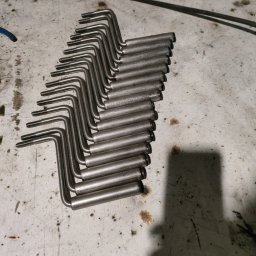 PREMIUMSTAL SEBASTIAN PRZYBYSZ - Rząd identycznych, surowych, metalowych prętów z gwintem, ułożonych równolegle na brudnym, białym stole warsztatowym, z widocznym kablem w tle.