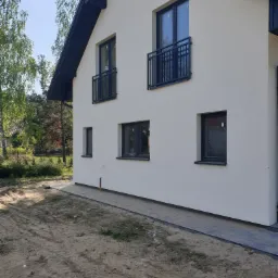 Nowoczesny dom jednorodzinny z jasną elewacją, ciemnymi oknami i balustradami balkonowymi, widok z zewnątrz na niezagospodarowany teren.