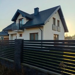 PREMIUMSTAL SEBASTIAN PRZYBYSZ - Nowoczesny dom z czarnym, poziomym ogrodzeniem panelowym na tle błękitnego nieba. Widoczne detale: betonowe słupki i podmurówka. Ujęcie z poziomu gruntu.