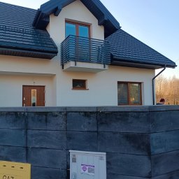 PREMIUMSTAL SEBASTIAN PRZYBYSZ - Nowoczesny dom jednorodzinny z balkonem i metalową balustradą w kolorze grafitowym. Widoczna elewacja w jasnym odcieniu i ciemna dachówka. Na pierwszym planie mur z szarej kostki.