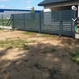 PREMIUMSTAL SEBASTIAN PRZYBYSZ - Nowoczesne, poziome ogrodzenie panelowe w kolorze grafitowym, z furtką, na tle domu z szarą dachówką i błękitnego nieba. Widoczne słupki i przęsła ogrodzenia.